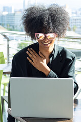 Feeling: Young non-binary person with an afro and sunglasses, wearing a black business suit, touching their chest excitedly while looking at their laptop.