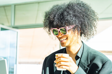 Happiness: Young non-binary person with an afro and sunglasses, wearing a black business suit. Holding a yellow coffee cup and smiling.