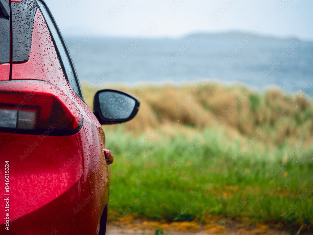 Wall mural a red car is parked on the side of a road next to a grassy field with tall grass on a dune and ocean