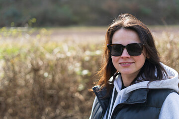 A smiling dark-haired girl in sunglasses stands in a park against the backdrop of tall reeds. The concept of unity between man and nature