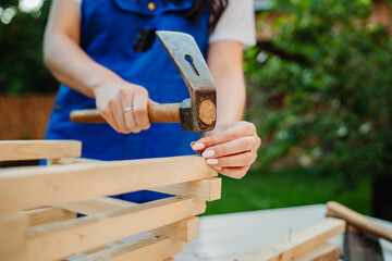 One young caucasian carpenter woman is using sawdust and hammer making things from wood and planks in her backyard during the day	