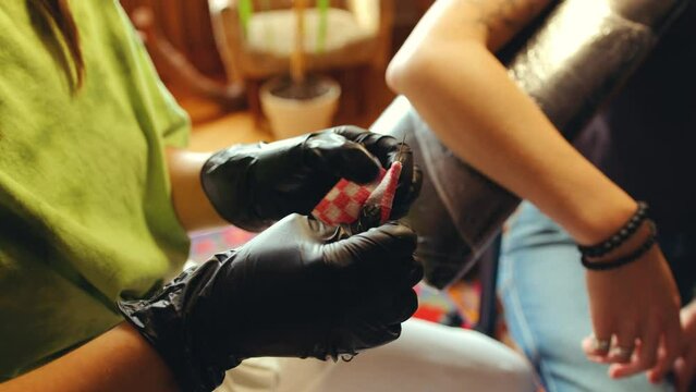 Hands of tattoo artist in sterile gloves preparing needle for tattooing arm of female client in salon. Close-up view