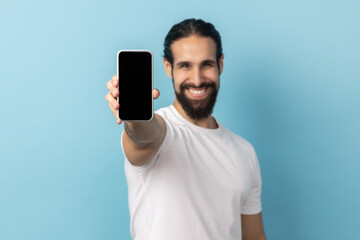 Portrait of positive man with beard wearing white T-shirt standing holding phone with empty display, looking at camera with toothy smile. Indoor studio shot isolated on blue background.
