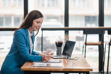 Smiling young woman in blue blazer working on laptop and smartphone at a wooden desk in a modern office with large windows.