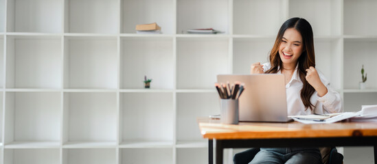 Excited young woman celebrating success while working on a laptop in a modern office with minimalist white shelves in the background.