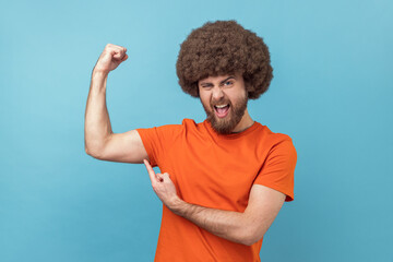Portrait of man with Afro hairstyle wearing orange T-shirt raises hand and indicates at biceps,...