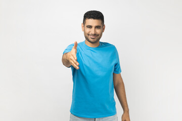 Man in blue T- shirt standing giving hand for handshake as sign of greeting, smiling widely and sincerely, looking very glad to see person he meeting. Indoor studio shot isolated on gray background.