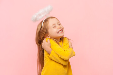 Portrait of cute smiling happy blonde little girl with nimb over head hugging herself with...