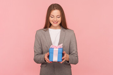 Portrait of delighted cheerful woman with brown hair holding blue wrapped present box, celebrating holiday, wearing business suit. Indoor studio shot isolated on pink background.