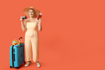 Mature female traveler with photo camera, passport and suitcase on orange background