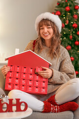 Young woman in Santa hat with Advent calendar sitting at home on Christmas eve
