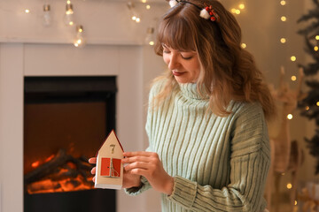 Young woman in reindeer horns with Advent calendar near fireplace at home on Christmas eve