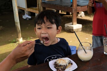  A young boy eagerly opens his mouth as he is fed a spoonful of dessert by an adult hand. His...