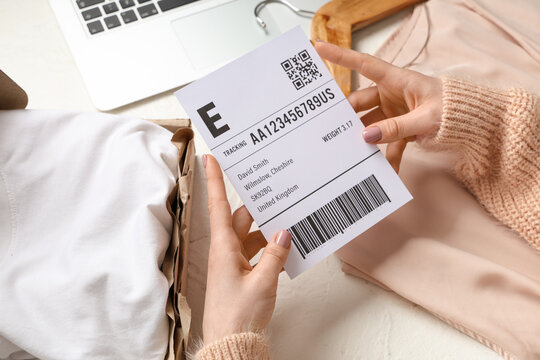 Female hands holding parcel label near open package, clothes and laptop on white table