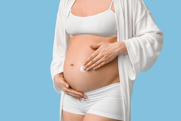 Young pregnant woman applying cream against stretch marks on blue background