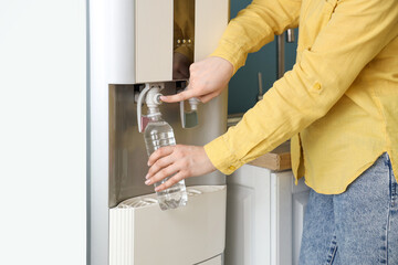 Woman filling plastic bottle with filtered water from cooler near blue wall