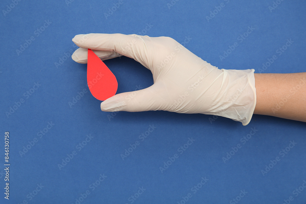 Wall mural female hand in rubber glove holding paper blood drop on blue background. donation blood concept. - Wall murals