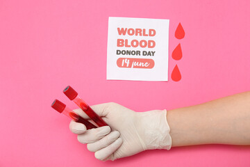 Female hand in rubber glove holding test tubes, paper drops and paper sheet with sign WORLD BLOOD DONOR DAY 14 JUNE on pink background