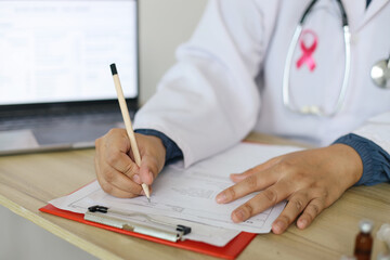 Doctor Wearing White Uniform With Pink Ribbon Sitting At Office And Writing On Medical Report For...