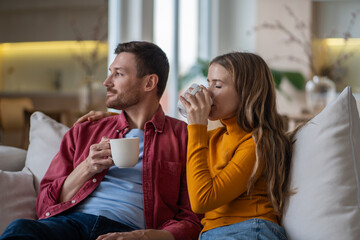 Relaxed family loving couple sits on couch with cup of coffee enjoy tranquility morning looking at window. Calm pleased wife woman drinking tea with husband man daydreaming, looking at window at home.