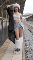 Pretty young Asian woman wears denim shorts and faux fur hat and boot cover on a train station platform