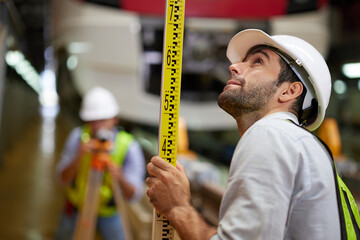Worker or engineer using theodolite surveying optical instrument and coworker holding large ruler...