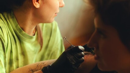 Young woman in gloves doing hand-poked tattoo with needle on arm of female client while working in tattoo studio