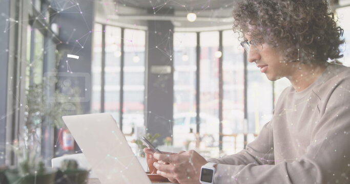 Young biracial man using smartphone while working on laptop in modern cafe - Powered by Adobe