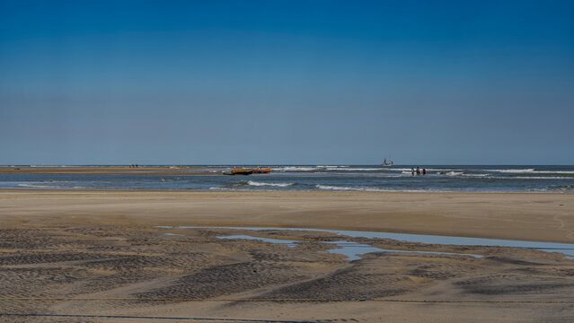 The low tide in the ocean. There are puddles of water on the exposed sand. Silhouettes of a team of fishermen in the surf, pulling out a fishing net. Pirogue boats in shallow water. A sailboat 