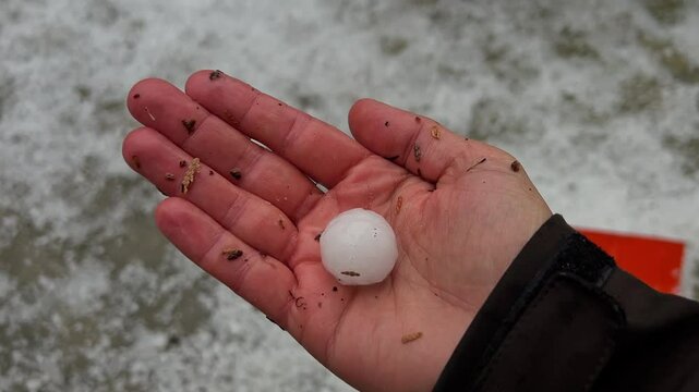 Top shot of a caucasian hand holding s single hailstone, hand dropping the ice sphere, camera focuses on the background showing a lot of hailstone laying in the driveway. shot in Wei&szlig;enhorn, Bavaria.