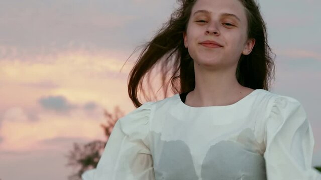 Happy teenage girl pouting and posing with raised hands against cloudy sky. Beautiful smiling female with long hair twirling during sunset. She is wearing white dress.