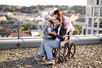 Family supporting man in wheelchair, using laptop together on urban rooftop. Embracing moments of togetherness and technology in an outdoor setting.