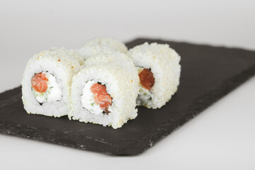 Close-up of four sushi rolls, coated in white sesame seeds, filled with salmon and cream cheese, served on a black slate plate