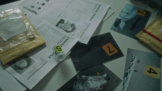 Close-up no people shot of criminal profile bulletins, numbered murder scene photos, phone, envelope with wedding ring in plastic bag, glasses, gun, laid out on table in interrogation room