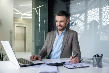 Fototapeta premium Confident businessman smiling while working on his laptop and taking notes in a sleek, modern office environment.