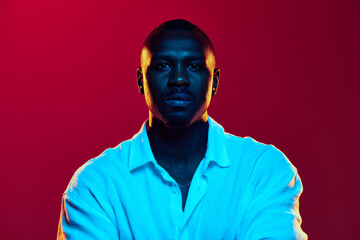 Portrait of African-American man in casual white shirt posing with serious face, showing confidence against red background in neon light. Concept of human emotions facial expression, fashion, business
