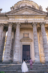 a beautiful couple of newlyweds are holding hands and walking towards an old building with columns and beautiful big doors.