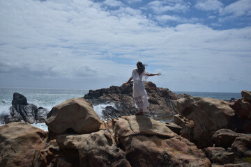 portrait of female model  standing wearing white goddess dress, dramatic natural landscape background of rocky ocean shoreline with stone clifftops. castle rock, Busselton, Western Australia