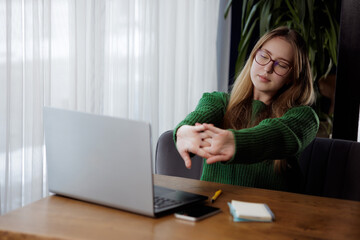 A woman is stretching her arms at a desk with a laptop