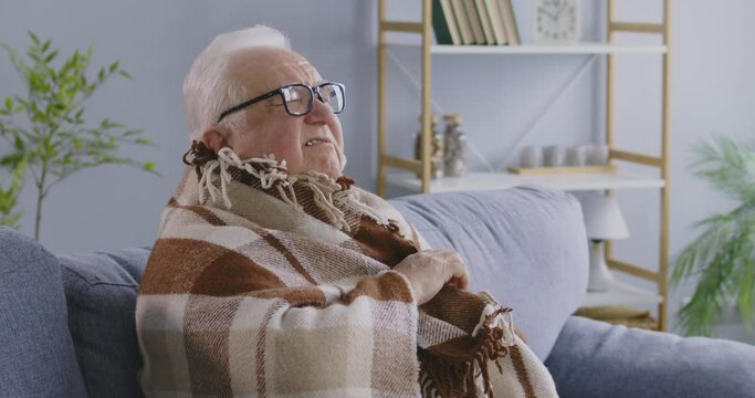 Senior gray-haired man trying to keep warm himself under a blanket sitting on sofa at home. Elderly retired person freezing in winter in chilly room with low temperature. 4k video.