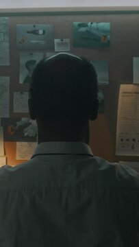 Rear vertical shot of nape of head of unrecognizable African American male detective standing front of crime map in office, examining photos of suspects and crime scene evidence, contemplating motive