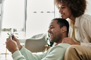 Joyful African American woman sitting near her husband with hearing aid and looking at phone.