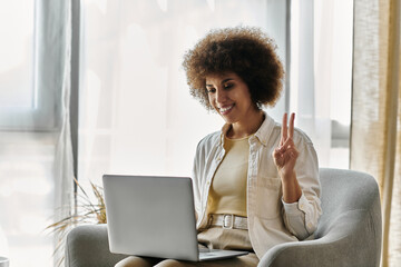 A woman sits in a chair, smiling as she uses sign language while looking at a laptop.