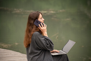 A woman is sitting on a dock with a laptop and a cell phone