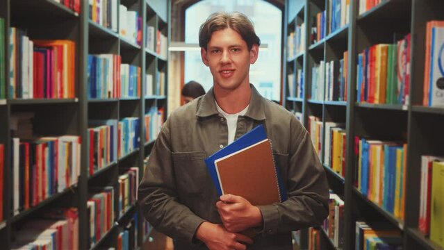 Zoom in Young man in gray jacket smiling and holding notebooks in library aisle, looking directly at camera. Colorful books and another student studying. Joy in learning and academic dedication