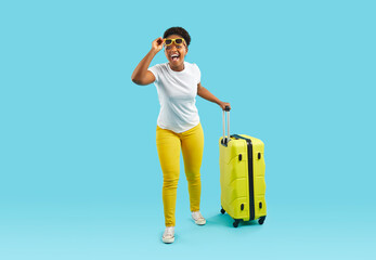 Vacation style dressed african american woman with hand luggage take off her sunglasses. Emotional movement forward, surprise and shock at what she saw. Isolated on blue background.