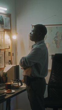 Medium vertical shot of African American police detective standing in front of crime map with photos of murder scene and potential suspects, scrutinizing evidence, while investigating gang murder