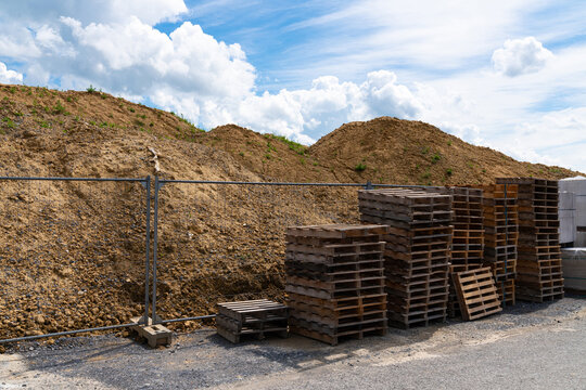 Wooden pallets stacked in front of a metal fence against the backdrop of large piles of earth under a light cloudy sky.
