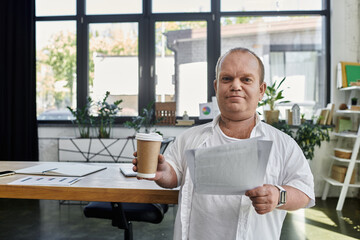 A man with inclusivity in a white shirt stands in an office setting, holding a cup of coffee and reviewing documents.