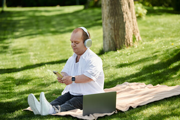 A man with inclusivity sits on a blanket in a park, listening to music with headphones and using his phone.
